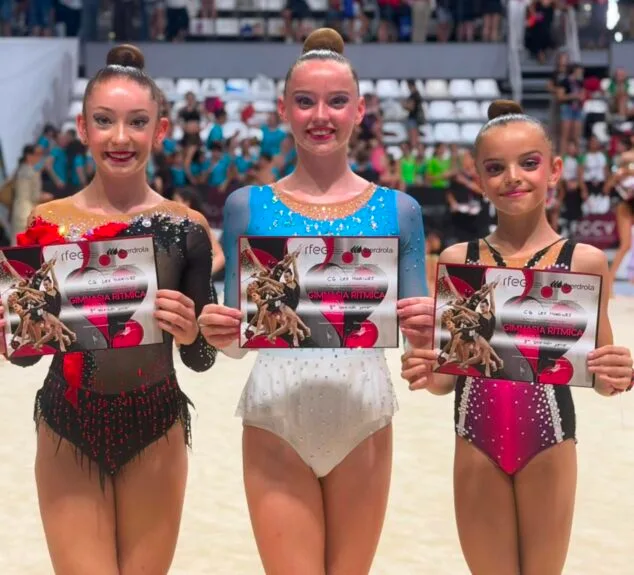 Imagen: Tres gimnastas del Club Les Marines sostienen diplomas en el interior de un pabellón deportivo tras el Campeonato de España.