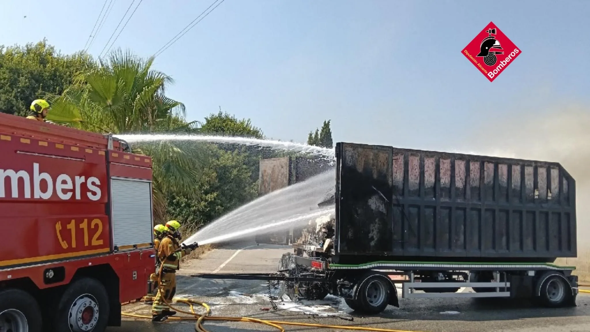 Bomberos extinguiendo con mangueras un incendio en el remolque abierto de un camión en una carretera.