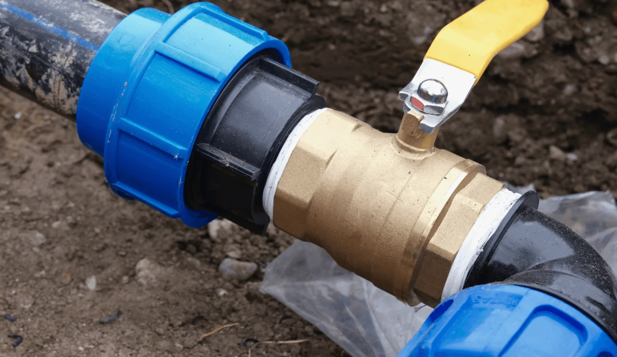 Primer plano de una llave de paso metálica con palanca amarilla instalada entre dos tuberías de agua potable sobre una zanja de tierra.