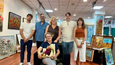 Calp nombrará Hija Predilecta a la pintora Lola Llinares en la gala del 9 d'Octubre 5 La pintora Lola Llinares sentada con flores y acompañada por un grupo en una galería de arte.