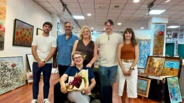 Calp nombrará Hija Predilecta a la pintora Lola Llinares en la gala del 9 d'Octubre 2 La pintora Lola Llinares sentada con flores y acompañada por un grupo en una galería de arte.