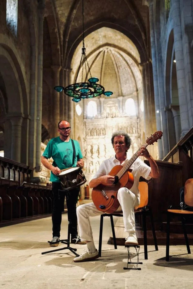 La Muixeranga de la Marina Alta protagoniza un histórico homenaje a Jaume I en el Monasterio de Poblet 2 Imagen: Dos músicos, uno sentado tocando la guitarra y otro de pie con un tambor, actúan en el interior de una iglesia de piedra con arcos góticos.