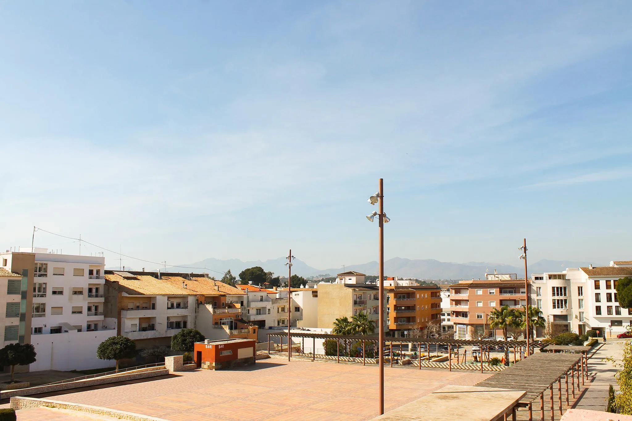 Vista de la Plaza de VI Centenari en Teulada, con suelo pavimentado y pocos árboles bajo un cielo soleado.