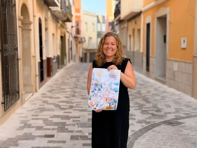 Imagen: Una mujer sonríe mientras sostiene el cartel del programa de fiestas de Santa María Magdalena 2026 en Benitatxell.