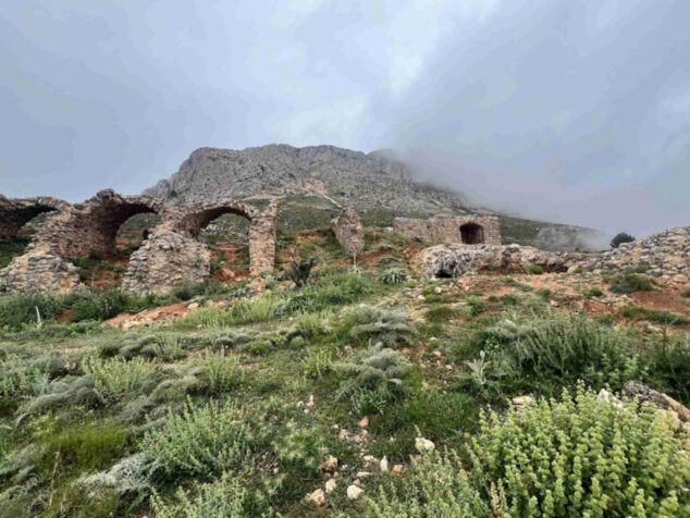 Imagen: Fotografía de las ruinas de piedra y arcos del Castillo de Bernia en la ladera de una montaña nublada.