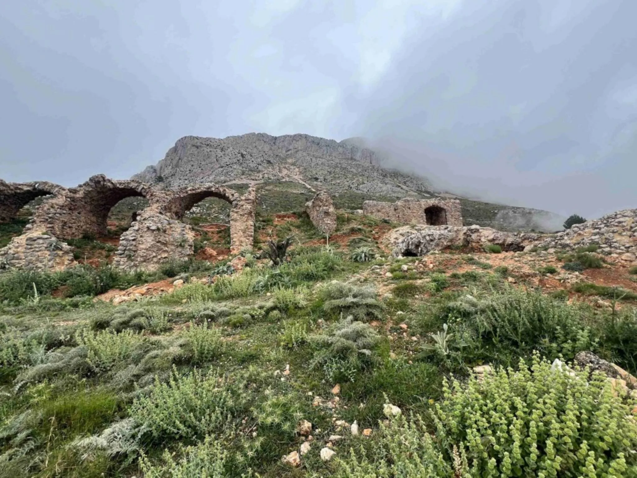 Fotografía de las ruinas de piedra y arcos del Castillo de Bernia en la ladera de una montaña nublada.