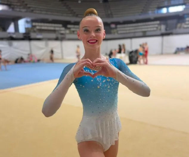 Imagen: La gimnasta Sophie O'Reilly posa sonriente haciendo un corazón con las manos en el pabellón de competición.