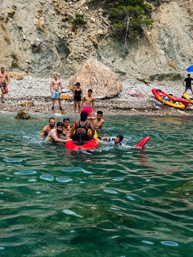 Rescatan a una mujer tras perder el conocimiento durante una excursión en kayak en Calp 1 Imagen: Varias personas en el agua socorren a una mujer subiéndola a un kayak rojo en una cala rocosa durante el rescate de la UVIM en Calp.