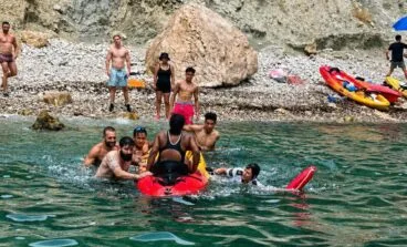 Varias personas en el agua socorren a una mujer subiéndola a un kayak rojo en una cala rocosa durante el rescate de la UVIM en Calp.