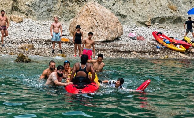Varias personas en el agua socorren a una mujer subiéndola a un kayak rojo en una cala rocosa durante el rescate de la UVIM en Calp.
