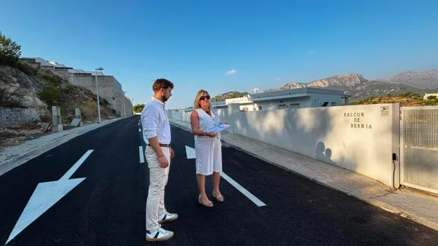 Imagen: La alcaldesa Ana Sala y el concejal Álex Coca observan la carretera recién asfaltada y señalizada de acceso a la urbanización Balcón de Bernia en Calp, con un muro y montañas al fondo.