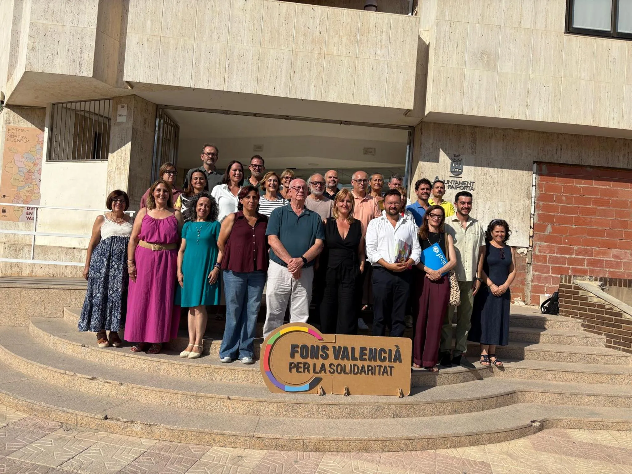 Grupo de personas posando en unas escaleras exteriores con un cartel de madera del Fons Valencià per la Solidaritat.