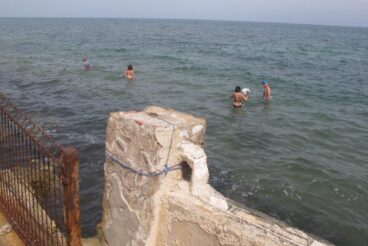 Bañistas en la costa de Dénia, cerca de una estructura y una valla.