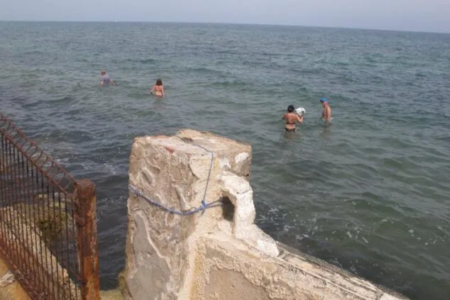Personas bañándose en el mar cerca de un muro y una valla metálica en la costa de Dénia.