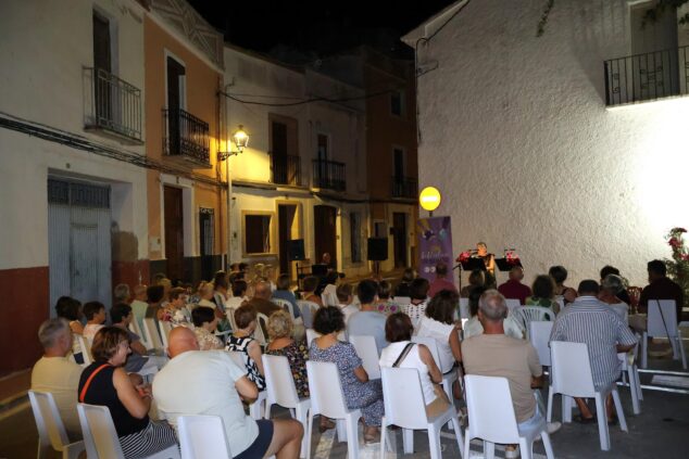 Imagen: Vista de una velada literaria nocturna al aire libre en una calle estrecha, con público sentado y una persona leyendo en un atril.