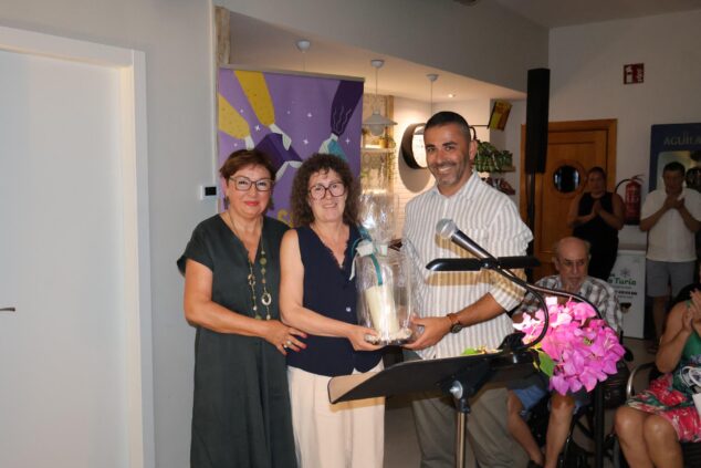 Benitatxell cierra con éxito el II Festival Literario 'Literatura a la fresca' 6 Dos mujeres entregan un regalo envuelto a un hombre en un acto interior con una pancarta al fondo, marcando el cierre del festival literario.