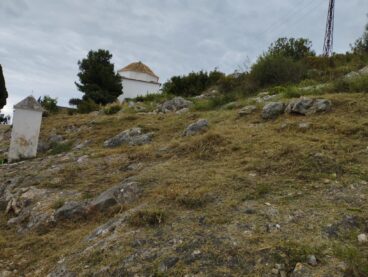 Pedreguer actúa contra incendios con un gran desbroce en dos zonas montañosas 2 Vista de una ladera rocosa y con vegetación desbrozada, con una pequeña capilla blanca con tejado de tejas en la parte superior, bajo un cielo nublado en Pedreguer.