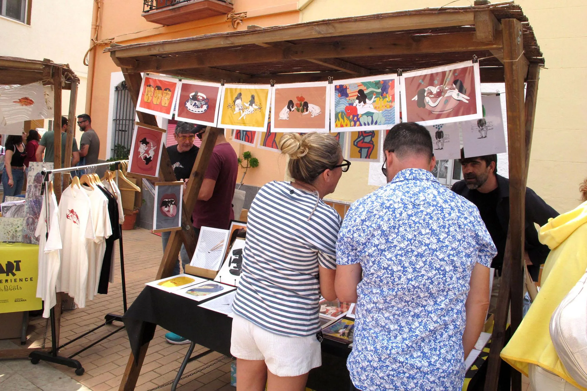 Visitantes mirando láminas ilustradas y camisetas en un puesto de arte al aire libre durante el Mercat d'Art.