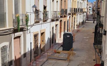 Las obras en la calle La Bassa de Gata de Gorgos se reanudan tras el hallazgo de los restos arqueológicos 3 Una calle en Gata de Gorgos en obras, con el suelo de tierra, vallas amarillas y un baño portátil al fondo.