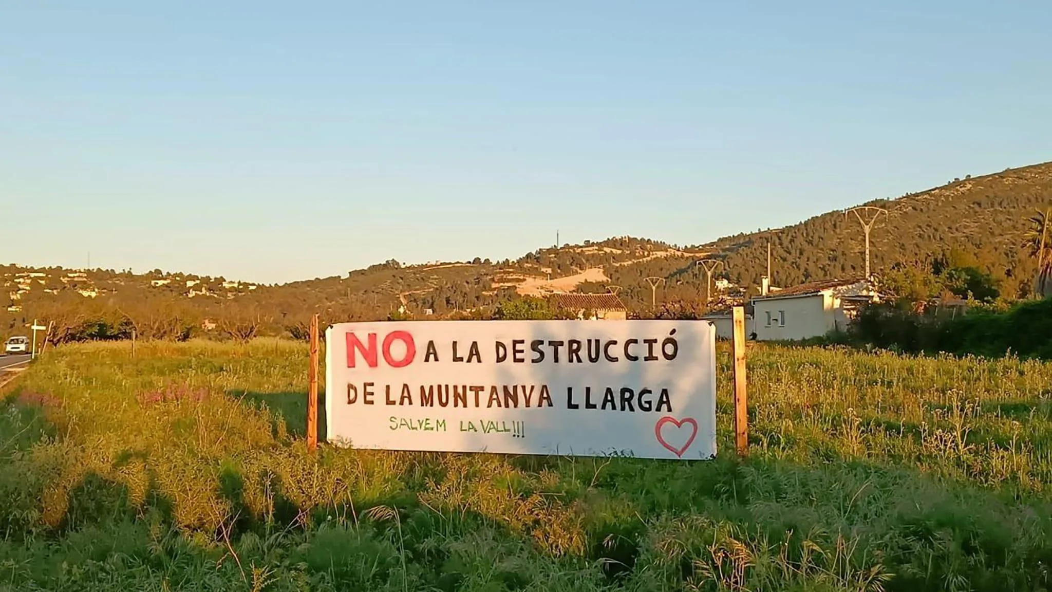 Pancarta blanca grande en un campo con el texto en rojo y negro: "NO A LA DESTRUCCIÓ DE LA MUNTANYA LLARGA. SALVEM LA VALL!!!" y un corazón dibujado.