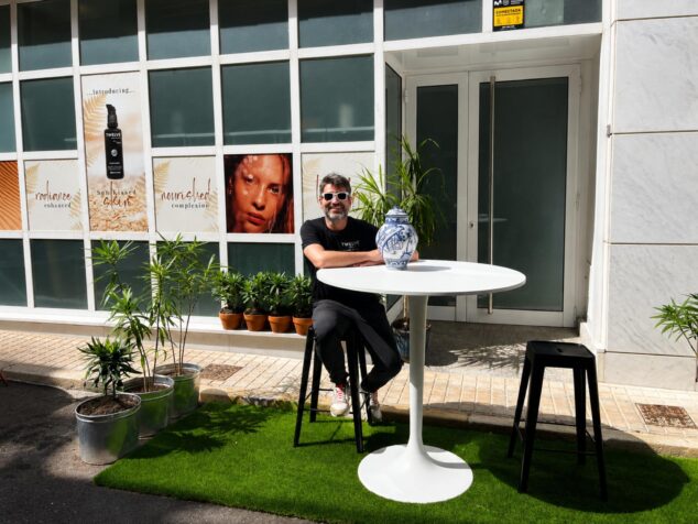«Cambiemos los aparcamientos por jardines»: la iniciativa del comercio local de Ondara 1 Imagen: Hombre sentado en una mesa en una plaza de aparcamiento transformada en pequeño jardín urbano con plantas y césped artificial frente a un comercio.