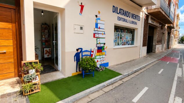 «Cambiemos los aparcamientos por jardines»: la iniciativa del comercio local de Ondara 2 Imagen: Exterior de la librería Dilatando Mentes en Ondara con un mural en la fachada y una pequeña instalación de jardín urbano con césped artificial, una silla azul y plantas frente a la entrada.