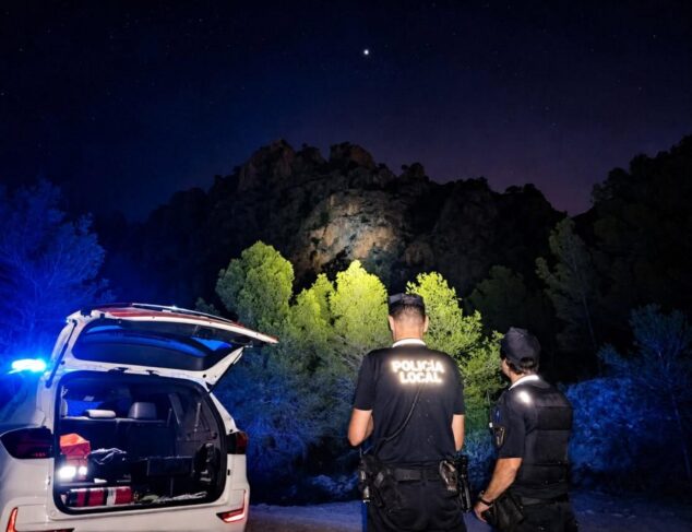 Imagen: Dos agentes de la Policía Local observan la montaña iluminada durante una búsqueda nocturna junto a un coche patrulla.