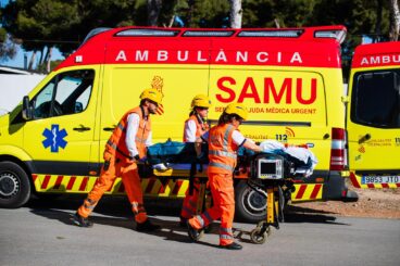 Tres miembros del equipo SAMU con cascos y uniformes naranjas empujan una camilla con un paciente hacia una ambulancia SAMU amarilla y roja.