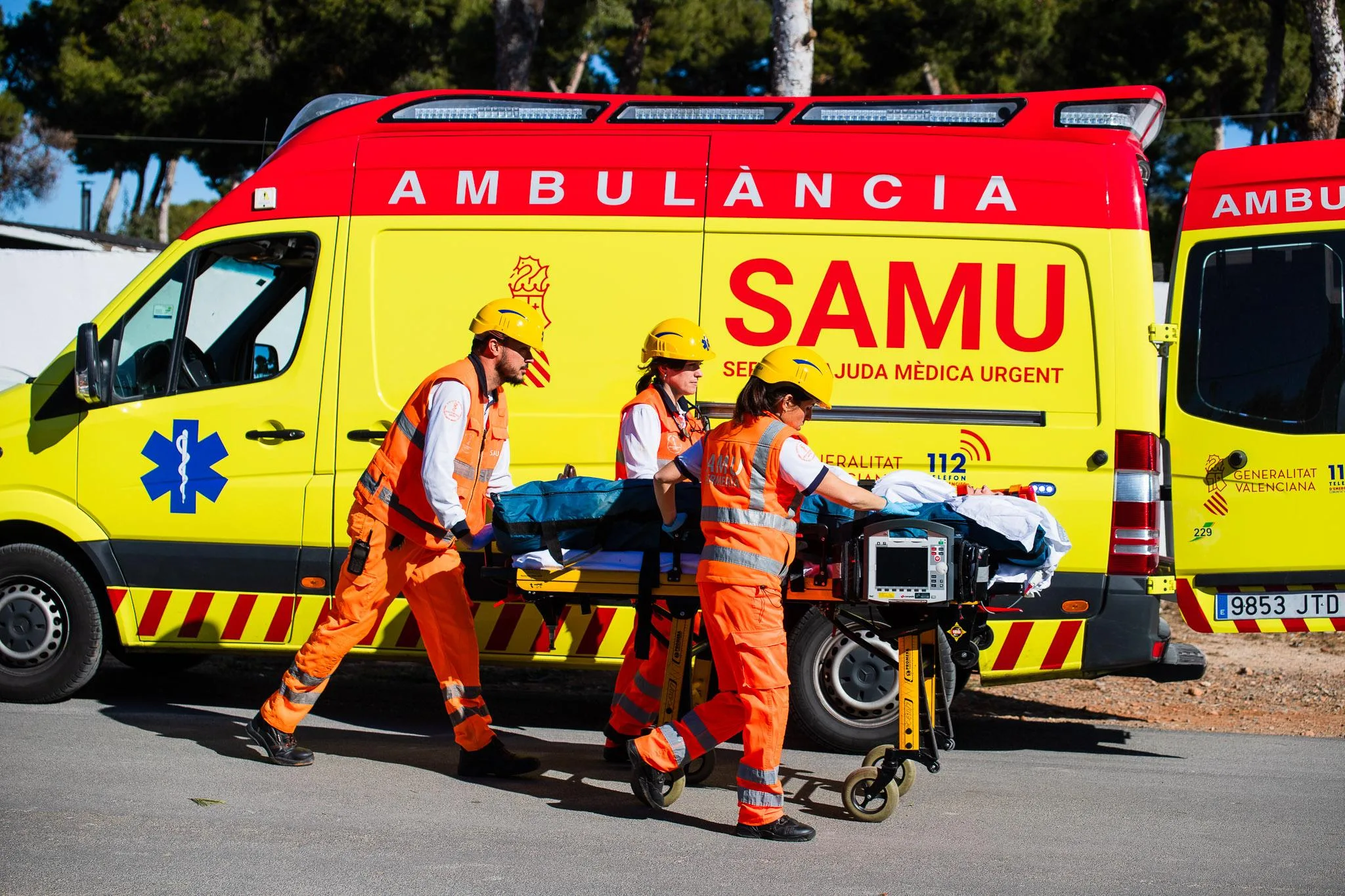 Tres miembros del equipo SAMU con cascos y uniformes naranjas empujan una camilla con un paciente hacia una ambulancia SAMU amarilla y roja.