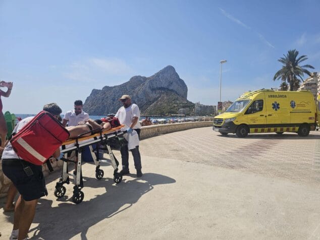 Calp pone a prueba su servicio de socorrismo con un rescate en la playa de la Fossa 3 Imagen: Varios socorristas y técnicos sanitarios trasladan a una persona en una camilla hacia una ambulancia amarilla aparcada en el paseo marítimo de la playa de la Fossa en Calp, con el peñón de Ifach visible al fondo.