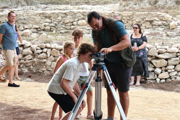 Imagen: Niños aprendiendo a usar un instrumento topográfico con un arqueólogo en un yacimiento arqueológico.