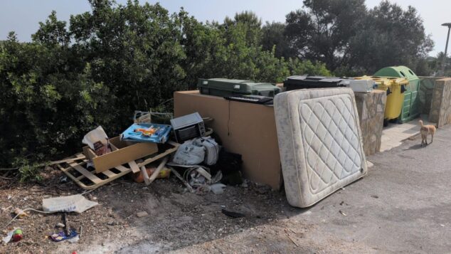 Imagen: Fotografía que muestra un colchón, un horno pequeño, maderas y bolsas de basura abandonados en el suelo junto a unos contenedores de reciclaje verdes y amarillos.