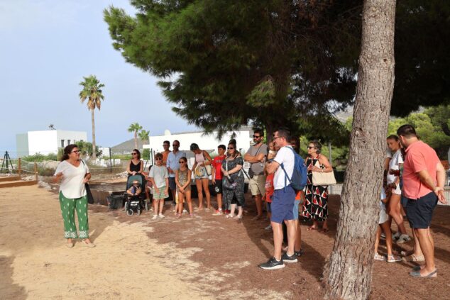 Imagen: Una mujer guía a un grupo de adultos y niños en un entorno natural con árboles durante una visita cultural al aire libre.