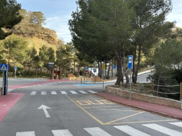 Zona de parada de autobús señalizada en el suelo del parking de acceso a la Cala Moraig, con vegetación y colinas al fondo.