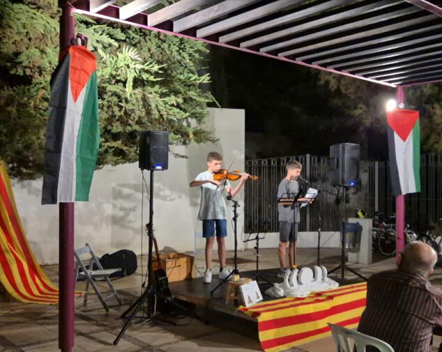 La Associació Cultural Ocell de Ondara rinde homenaje a Vicent Andrés Estellés con poesía y solidaridad con el pueblo palestino 1 Imagen: Dos jóvenes sobre un escenario nocturno al aire libre, uno tocando el violín y otro leyendo, con banderas de Palestina y la Comunidad Valenciana.