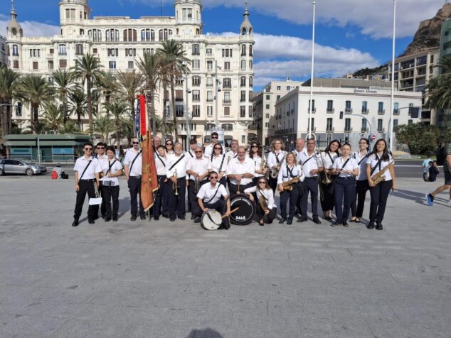 Imagen: Fotografía de grupo de los miembros de la Asociación Musical Jalonense de Xaló con sus instrumentos y estandarte en una plaza.