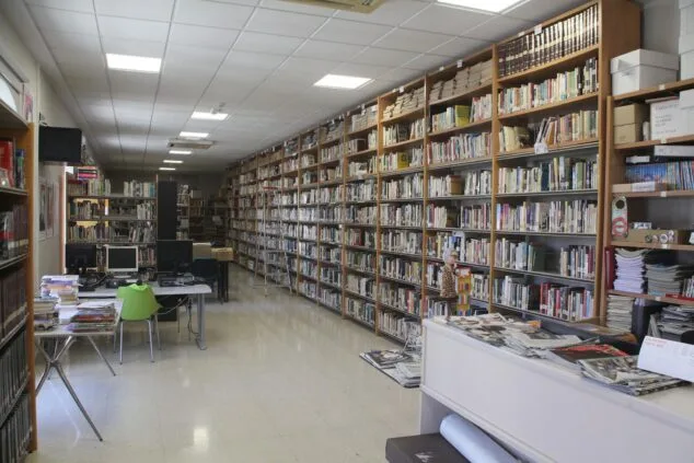 Vista del interior de la biblioteca municipal de Pedreguer con estanterías llenas de libros, mesas de estudio con ordenadores y un mostrador de préstamo.