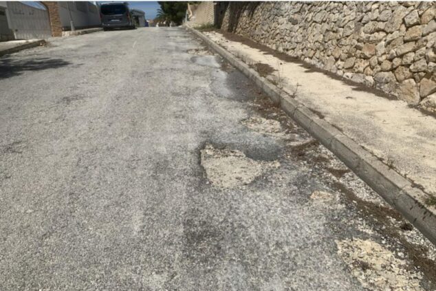 Imagen: Primer plano de una calle asfaltada en Calp mostrando baches profundos y deterioro severo junto a la acera y un muro de piedra.