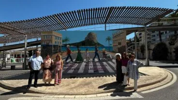 Grupo de seis personas posando frente a un mural grande y colorido sobre los derechos de las mujeres en una estación de autobuses al aire libre en Calp, con el Peñón de Ifach al fondo.