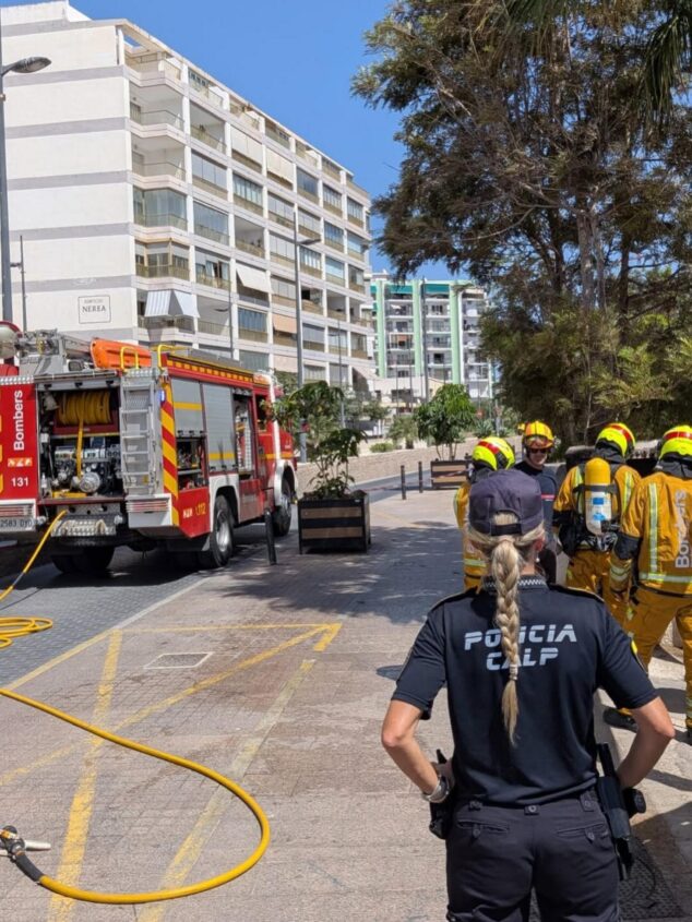 Un contador eléctrico se incendia en Calp y afecta a la instalación de varios edificios 2 Imagen: Agente de Policía Local de Calp de espaldas observando a bomberos trabajar junto a un camión de bomberos en una calle.