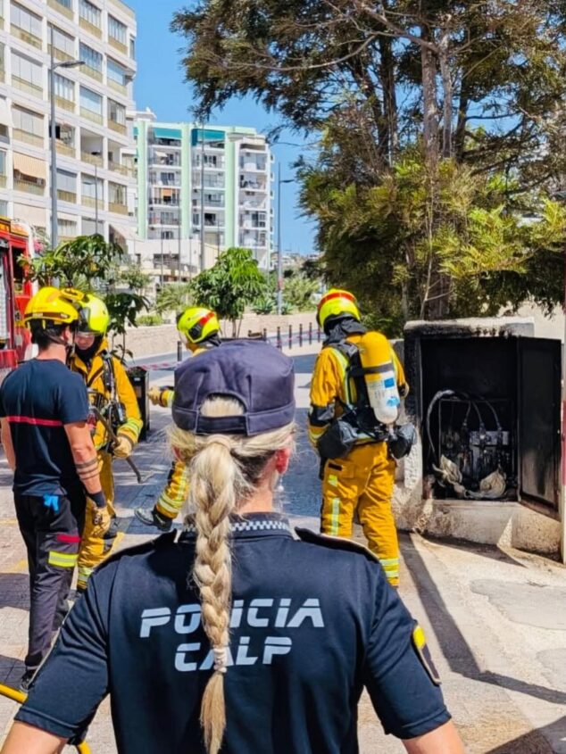 Un contador eléctrico se incendia en Calp y afecta a la instalación de varios edificios 1 Imagen: Agente de Policía Local de Calp de espaldas mirando a varios bomberos con equipos de protección trabajando en un cuadro eléctrico quemado abierto en una acera.