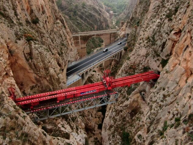 Imagen: Fotografía aérea de una gran estructura metálica roja de construcción sobre el barranco del Mascarat, con un nuevo puente debajo y un antiguo puente de piedra al fondo.