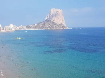 Vista panorámica de una playa de Calp con bañistas en el agua azul turquesa y el Peñón de Ifach al fondo.