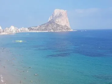 Vista panorámica de una playa de Calp con bañistas en el agua azul turquesa y el Peñón de Ifach al fondo.