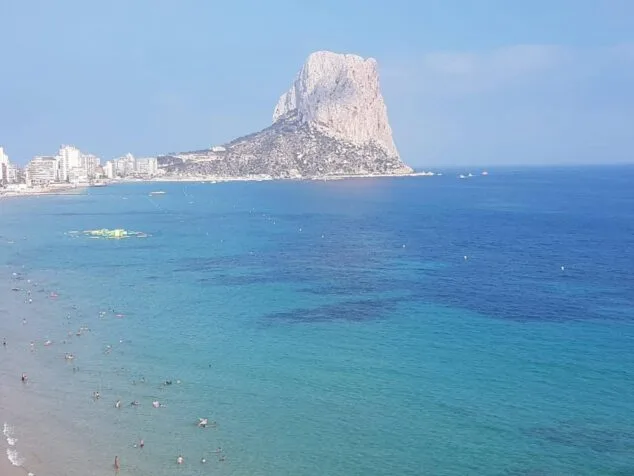 Vista panorámica de una playa de Calp con bañistas en el agua azul turquesa y el Peñón de Ifach al fondo.
