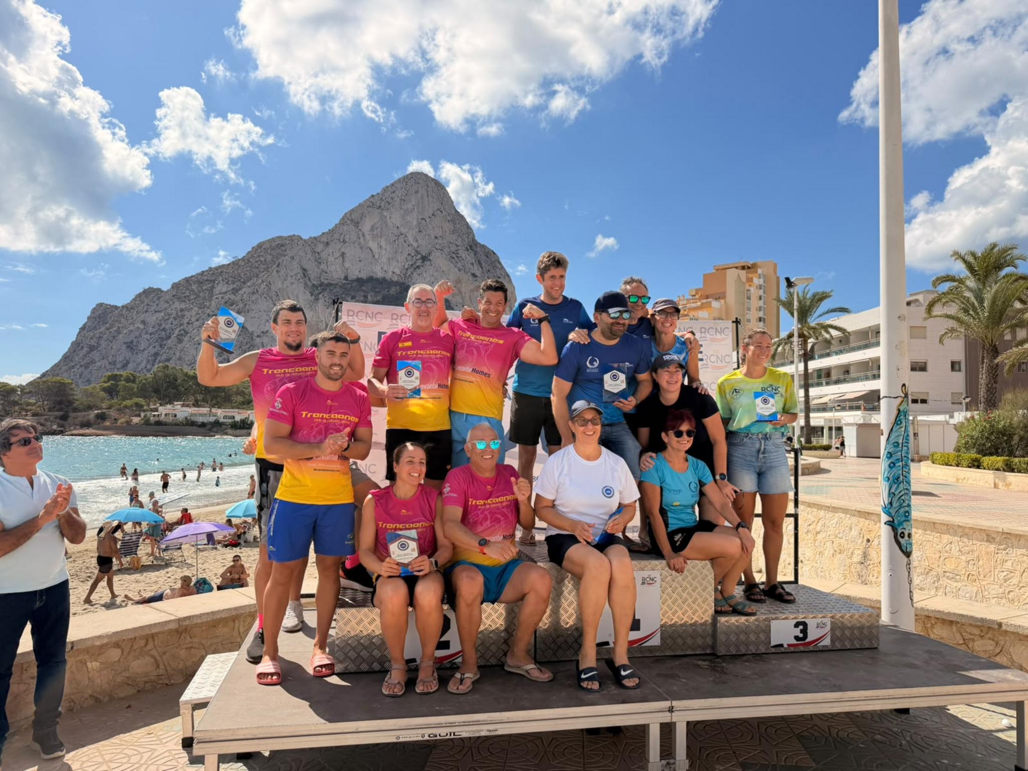 Grupo de nadadores del Club Trencaones en el podio de entrega de premios en la playa de Calpe con el Peñón de Ifach detrás.