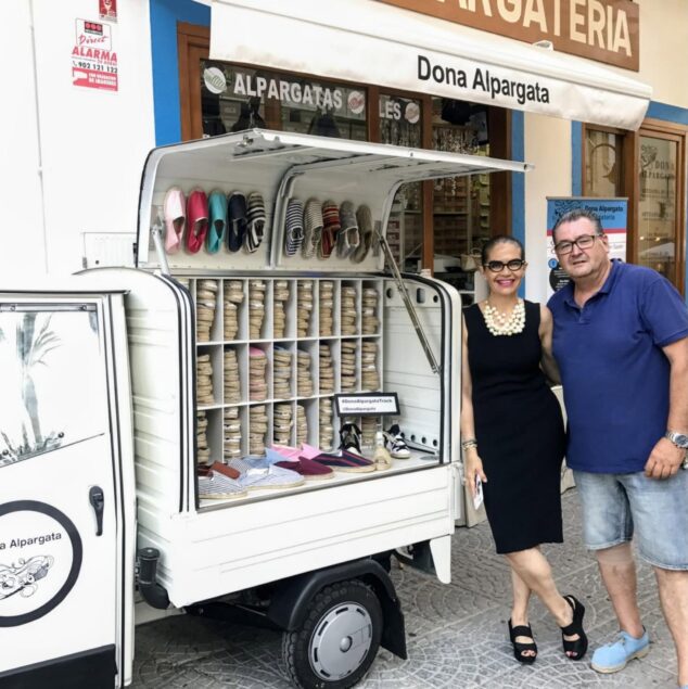 Una pareja de pie junto a un vehículo blanco que exhibe alpargatas frente a la tienda Dona Alpargata.
