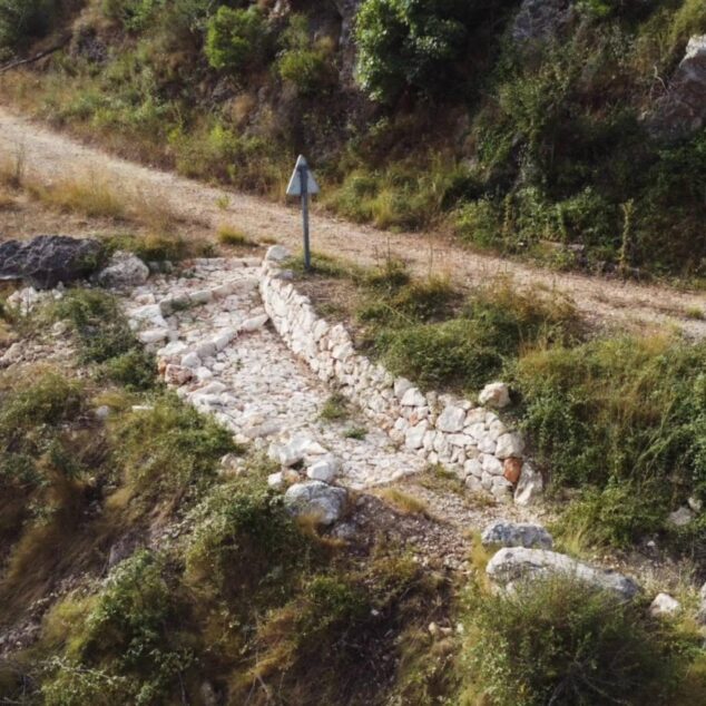 Ganadería extensiva y pedra en sec: la estrategia que se desarrolla en la Vall d’Ebo para cuidar el territorio 3 Imagen: Vista de una estructura de contención de piedra en seco en construcción, parte de un camino rural en un entorno montañoso y seco con vegetación.