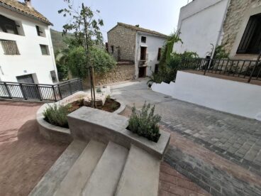 Fotografía de la Plaça de Baix de Benirrama, en La Vall de Gallinera, mostrando las nuevas escaleras de hormigón, jardineras y el pavimento renovado tras las obras de remodelación.