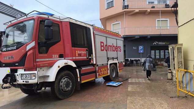 Imagen: Camión rojo de bomberos aparcado en una calle mojada con una persona caminando y un edificio al fondo.